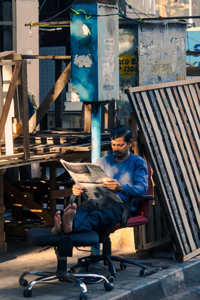 services-05 Casual scene of a man reading a newspaper on a chair in an urban Mumbai street.